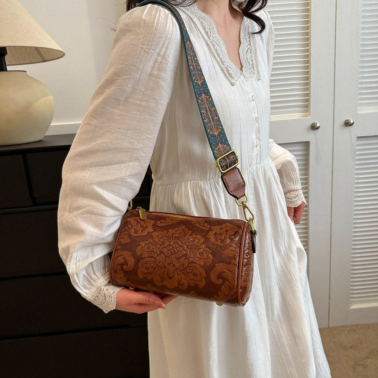Donna con abito bianco indossa borsa a tracolla in pelle marrone con motivo floreale. Accessorio moda elegante, perfetto per look casual chic.