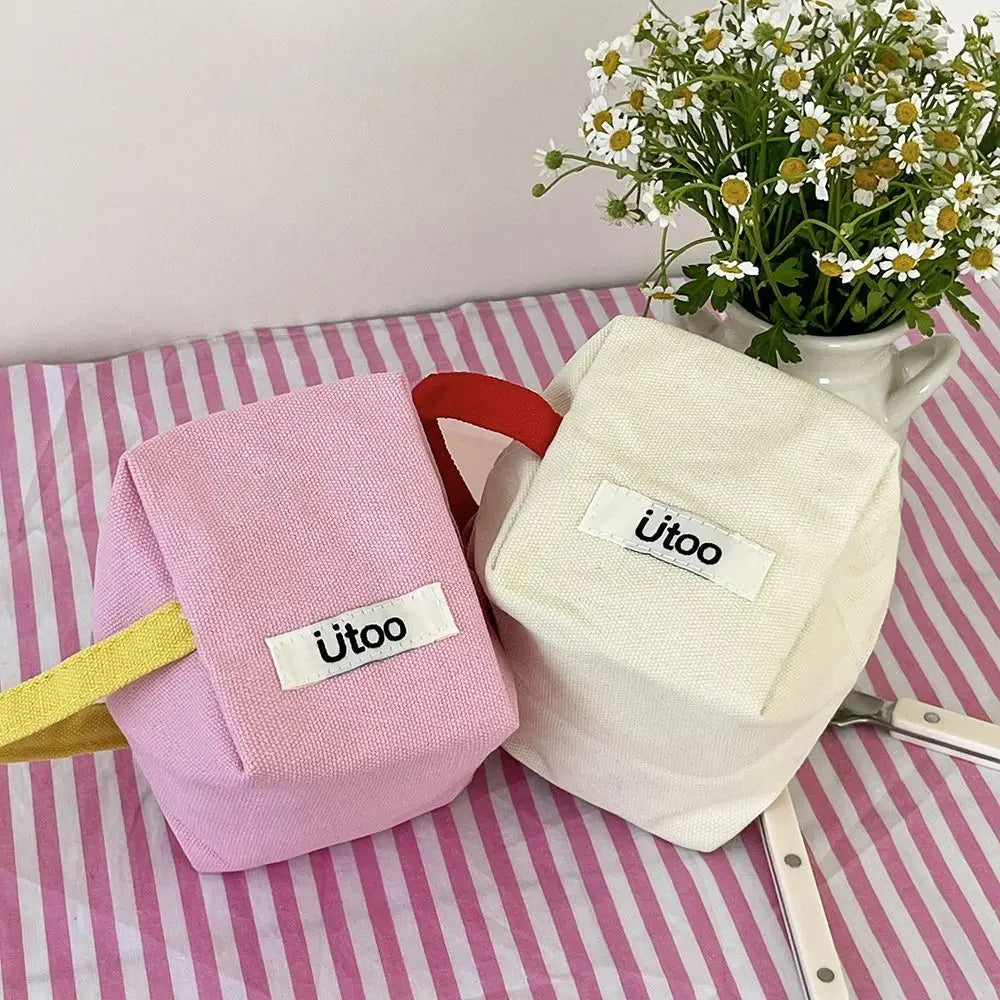 Due astucci in tessuto rosa e bianco con etichetta "Ütoo" su tovaglia a righe rosa, accanto a vaso di fiori bianchi. Accessori scuola eleganti.