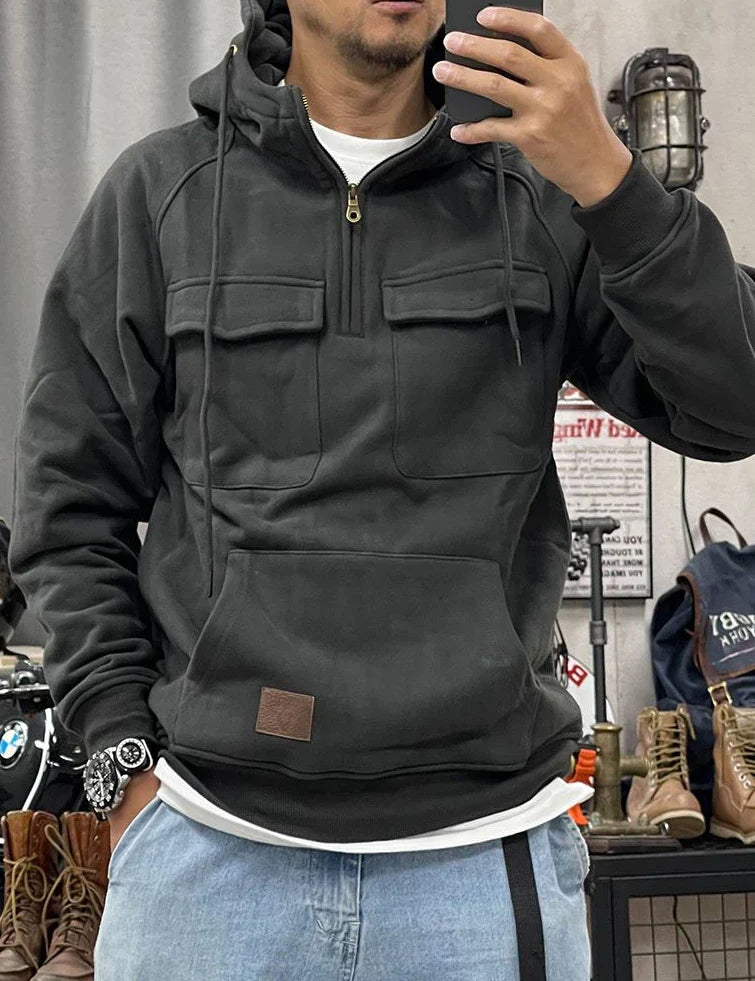 Uomo con felpa nera con cappuccio e tasche frontali, jeans chiari, orologio al polso, scatta selfie in negozio di abbigliamento casual.