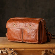 Borsa a tracolla in pelle marrone vintage su tavolo di legno, design con doppia tasca frontale, ideale per uomo e donna, accessorio moda elegante.