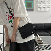 Borsa a tracolla nera in pelle, indossata da una persona con t-shirt bianca e pantaloni scuri, in un salotto moderno. Accessorio moda elegante.