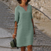 Donna con abito verde salvia, maniche a tre quarti, scollo a V, cammina su strada acciottolata. Accessori: cappello, bracciali. Moda casual elegante.