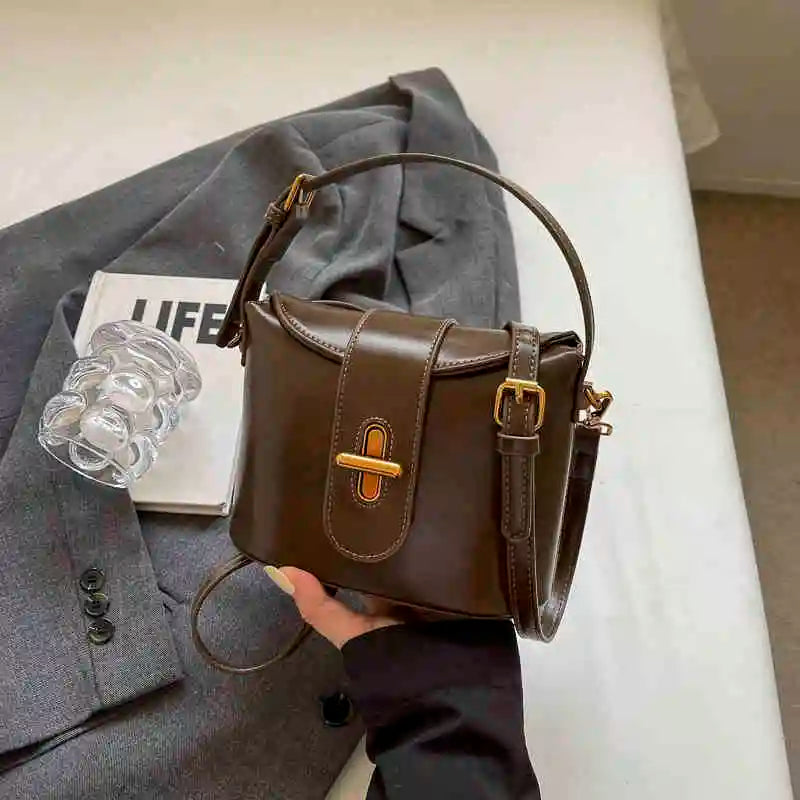 Borsa a tracolla in pelle marrone con fibbia dorata, posata su giacca grigia. Accessorio moda elegante, perfetto per look autunnali.