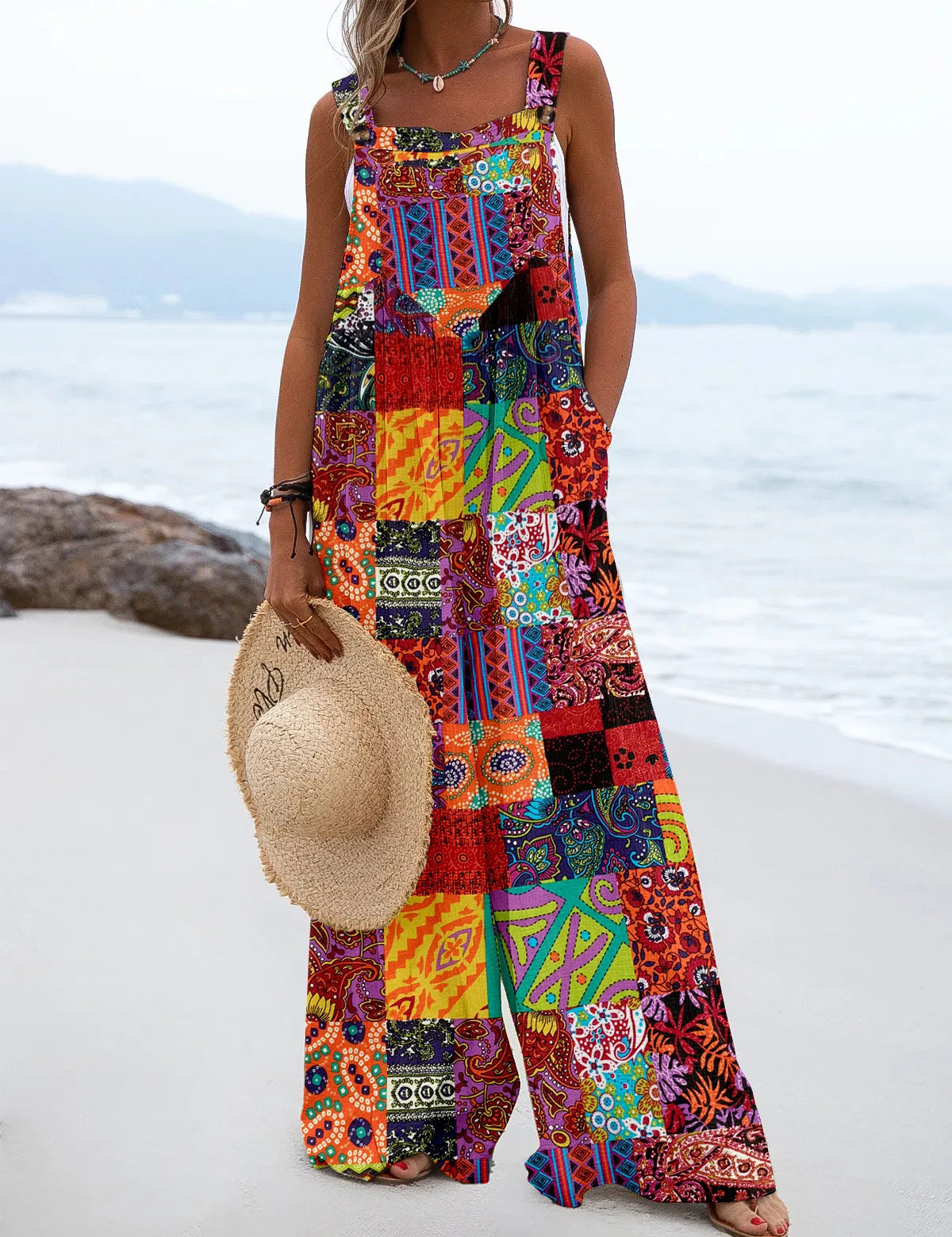 Donna in spiaggia con tuta lunga patchwork colorata, cappello di paglia in mano. Moda estiva, abbigliamento boho chic, abito casual vivace.