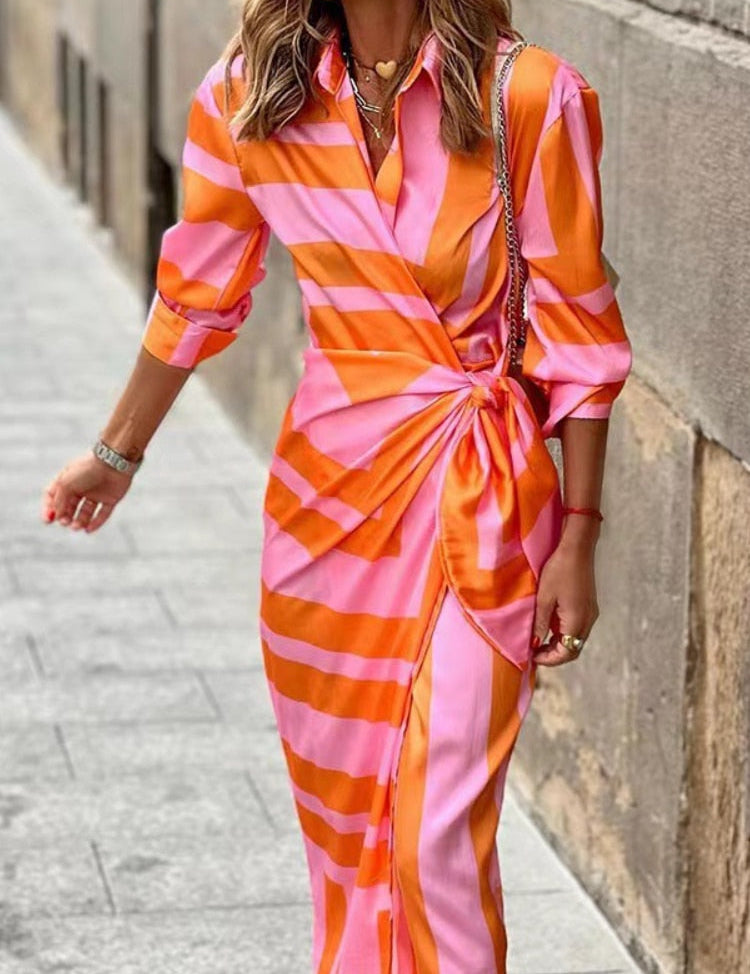 Abito lungo a righe rosa e arancioni, maniche lunghe, scollo a V, nodo in vita, indossato da donna su strada urbana. Moda elegante e colorata.