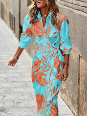 Abito lungo floreale azzurro e arancione, maniche lunghe, scollo a V, donna sorridente, strada cittadina. Moda estiva elegante.