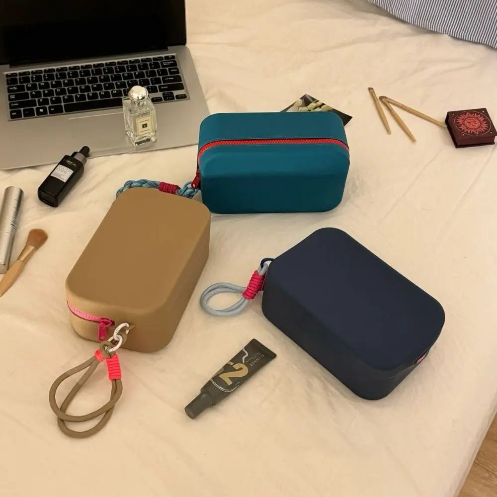 Tre astucci colorati su un letto accanto a un laptop, profumo e trucchi. Astucci in silicone con cerniera, ideali per cosmetici e accessori.