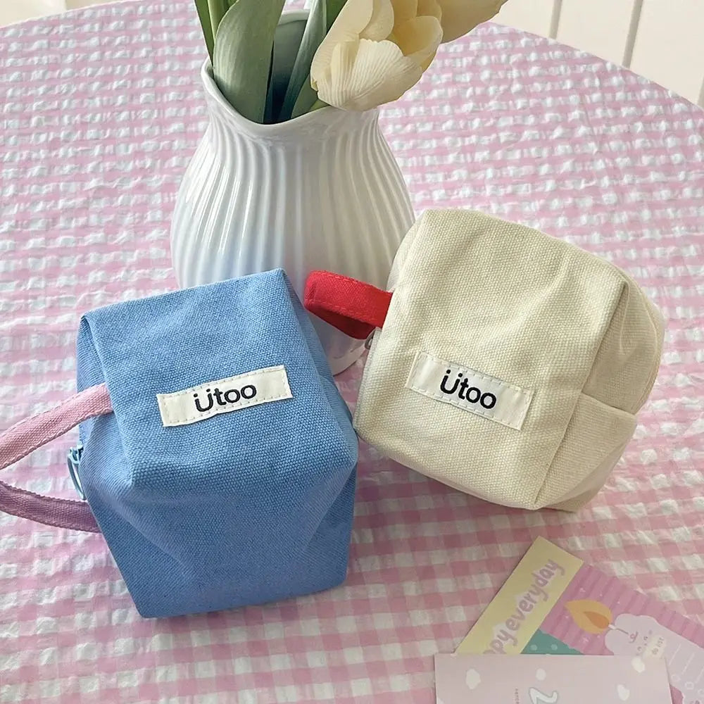 Due astucci cubici in tessuto, uno blu e uno beige, con etichetta "jitoo", su tavolo a quadretti rosa, accanto a vaso bianco con fiori.