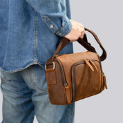 Uomo con camicia di jeans che tiene una borsa a tracolla in pelle marrone, design vintage, tasche con cerniera, moda casual, accessorio maschile.