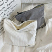 Due bustine in tessuto a coste, una bianca e una grigia, su un letto bianco. Design minimalista, ideale per accessori moda o cosmetici.