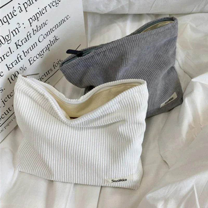 Due bustine in tessuto a coste, una bianca e una grigia, su un letto bianco. Design minimalista, ideale per accessori moda o cosmetici.