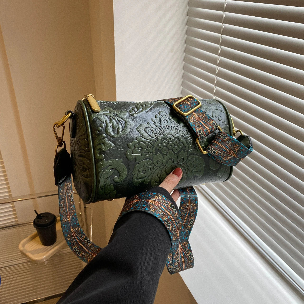 Borsa a tracolla cilindrica verde con motivo floreale in rilievo, fibbia dorata e tracolla decorata. Accessorio moda elegante e versatile.
