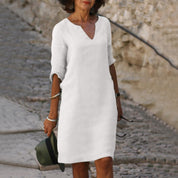 Donna con abito bianco casual, maniche a tre quarti, scollo a V, cammina su strada acciottolata. Stile estivo elegante e minimalista.