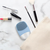 Dispositivo di pulizia viso blu su marmo bianco, circondato da pennelli trucco e rossetto rosso. Accessori bellezza, cura della pelle, cosmetici.