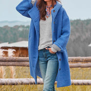 Donna con cappotto blu in lana, maglione grigio, jeans, e sciarpa colorata, in un ambiente rurale. Moda autunnale, abbigliamento casual elegante.