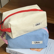 Due astucci in tessuto, uno beige con cerniera rossa e uno azzurro, su un libro. Ideali per scuola o viaggio. Marca Utoo.
