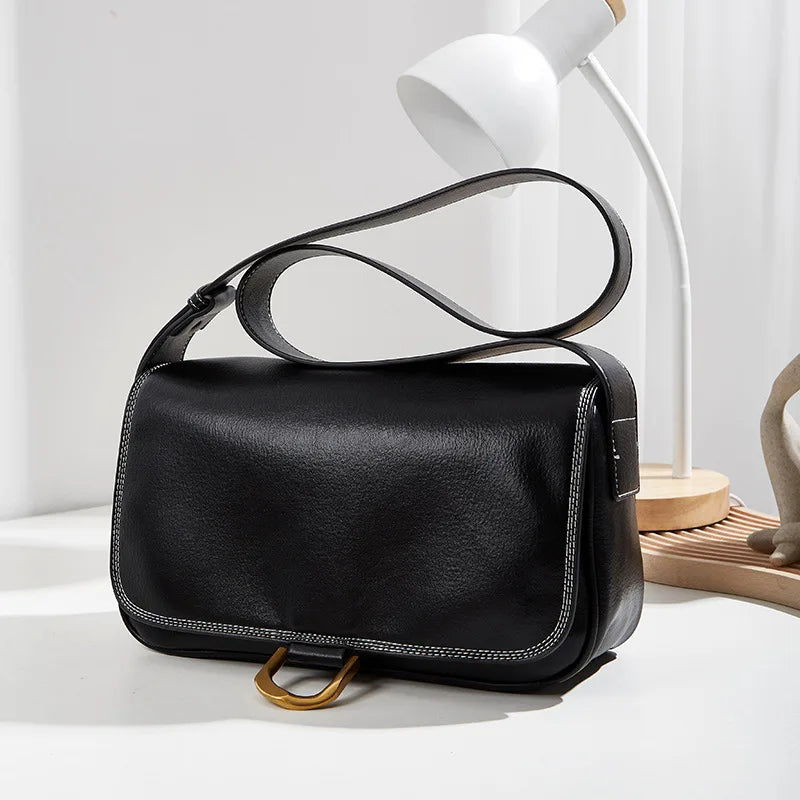 Borsa a tracolla in pelle nera su tavolo bianco, design elegante con cuciture a contrasto, lampada moderna sullo sfondo. Accessorio moda donna.