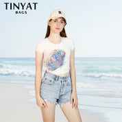 Giovane donna in spiaggia con marsupio olografico TINYAT, cappellino bianco e shorts di jeans. Accessorio moda estivo, stile casual.