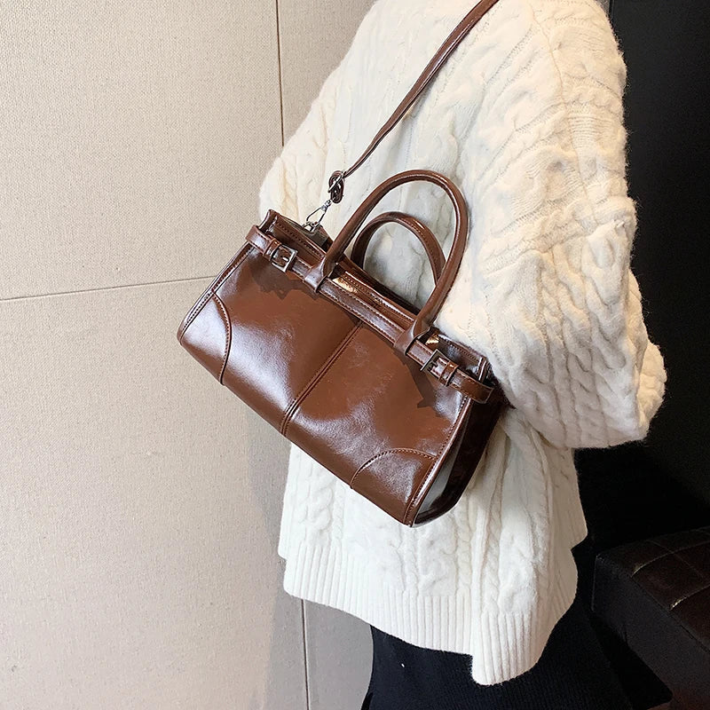 Borsa a mano in pelle marrone lucida su sfondo bianco, indossata da una persona con maglione bianco. Elegante accessorio moda autunno inverno.
