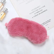 Maschera per dormire in pelliccia sintetica rosa, morbida e confortevole, su sfondo bianco con flaconi di profumo. Accessorio relax e benessere.