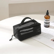 Borsa organizer nera in rete con cerniera su tavolo bianco, accanto a bottiglia di siero marrone. Accessori da viaggio, cosmetici, praticità.