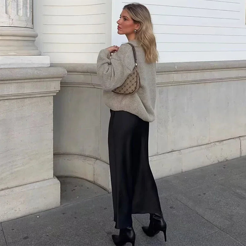 Donna con maglione beige, gonna lunga nera e borsa a tracolla marrone, cammina su marciapiede urbano. Scarpe nere con tacco alto. Moda autunnale elegante.