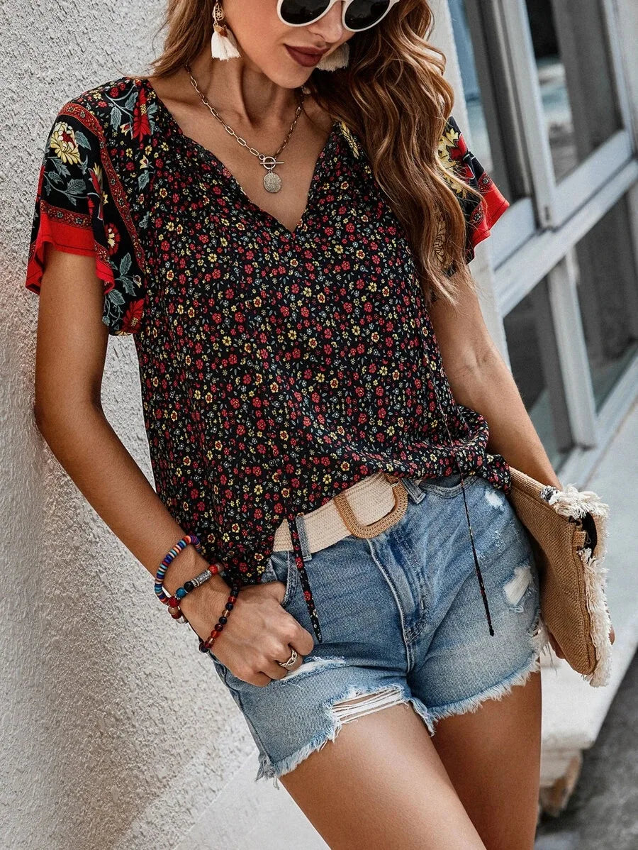Donna con occhiali da sole indossa blusa floreale nera e rossa, shorts di jeans, accessori colorati, borsa beige. Moda estiva casual femminile.