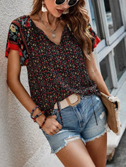 Donna con occhiali da sole indossa blusa floreale nera e rossa, shorts di jeans, accessori colorati, borsa beige. Moda estiva casual femminile.
