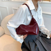 Donna con camicia bianca e borsa a spalla in pelle lucida bordeaux, seduta su poltrona beige. Eleganza, moda femminile, accessori di tendenza.