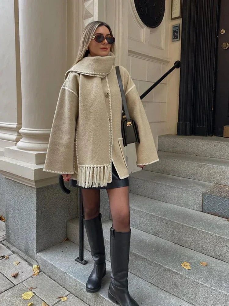 Donna con cappotto beige oversize e sciarpa abbinata, occhiali da sole, borsa a tracolla nera, stivali alti neri, su gradini di pietra. Moda autunnale elegante.