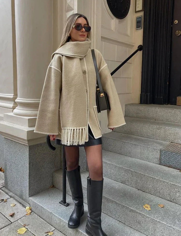 Donna con cappotto beige oversize e sciarpa abbinata, occhiali da sole, borsa a tracolla nera, stivali alti neri, su gradini di pietra. Moda autunnale elegante.