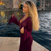 Donna bionda in abito viola su molo, sfondo di case e mare al tramonto. Moda elegante, atmosfera serale, località costiera italiana.