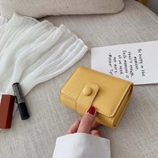 Portafoglio giallo in pelle su divano beige, accanto a rossetto nero e libro. Design compatto, chiusura a bottone. Accessorio moda elegante.
