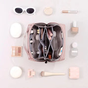 Borsa trucco rosa aperta con scomparti organizzati, circondata da cosmetici: occhiali da sole, ombretti, pennello, rossetto. Accessori beauty.