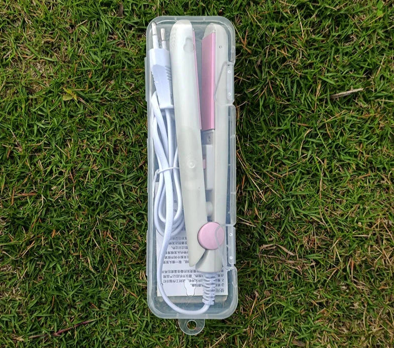 Piastra per capelli portatile bianca e rosa in custodia trasparente su erba verde, con cavo di alimentazione. Accessorio styling compatto e leggero.
