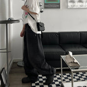 Utente in salotto moderno con divano nero, indossa jeans larghi neri, t-shirt bianca e borsa a tracolla nera. Arredamento minimalista.