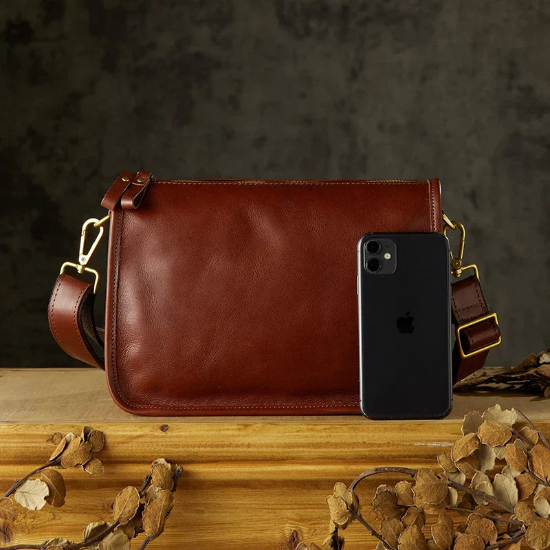Borsa a tracolla in pelle marrone con fibbie dorate, affiancata da uno smartphone nero. Elegante accessorio moda per uomo e donna.