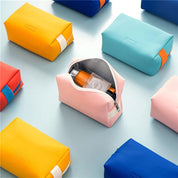 Astuccio da viaggio rosa aperto con prodotti cosmetici, circondato da astucci colorati su sfondo azzurro. Accessori organizzazione viaggio.