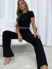 Donna con completo nero elegante, top corto e pantaloni a zampa, seduta su divano bianco. Moda femminile, abbigliamento casual chic, tendenze 2023.
