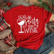 Maglietta rossa natalizia con scritta "Dear Santa Just Bring Wine", decorata con bicchiere di vino e fiocchi di neve, su sfondo di legno e decorazioni festive.