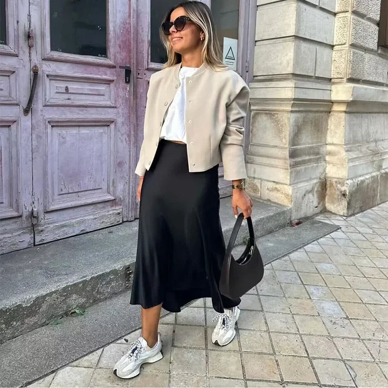 Donna con giacca beige, gonna nera e sneakers bianche, borsa nera a mano, occhiali da sole, posa davanti a porta antica. Moda casual elegante.