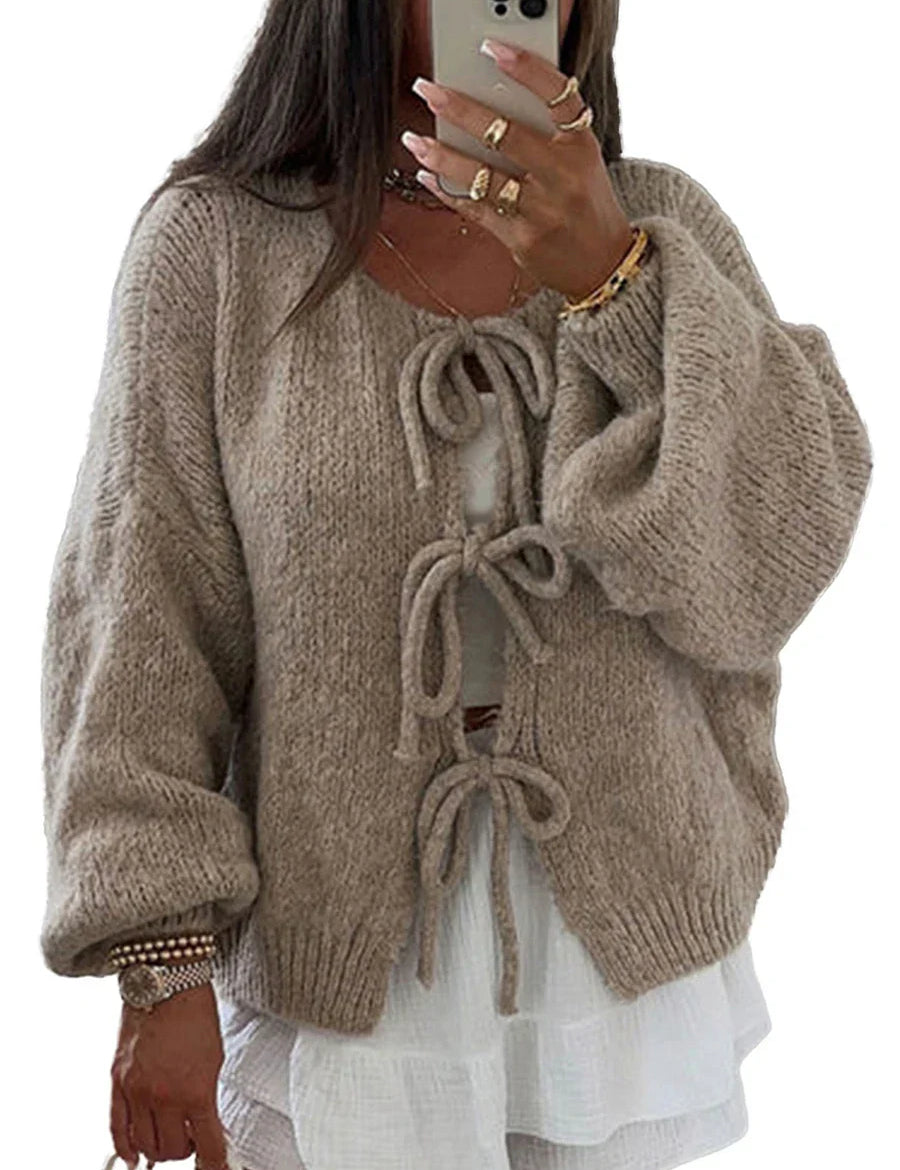 Donna con cardigan beige in maglia con fiocchi, maniche lunghe e ampie, sopra abito bianco. Moda autunnale, abbigliamento casual elegante.
