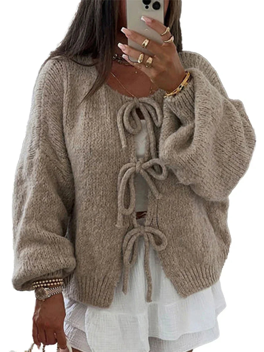 Donna con cardigan beige in maglia con fiocchi, maniche lunghe e ampie, sopra un abito bianco. Stile casual chic, moda autunnale.