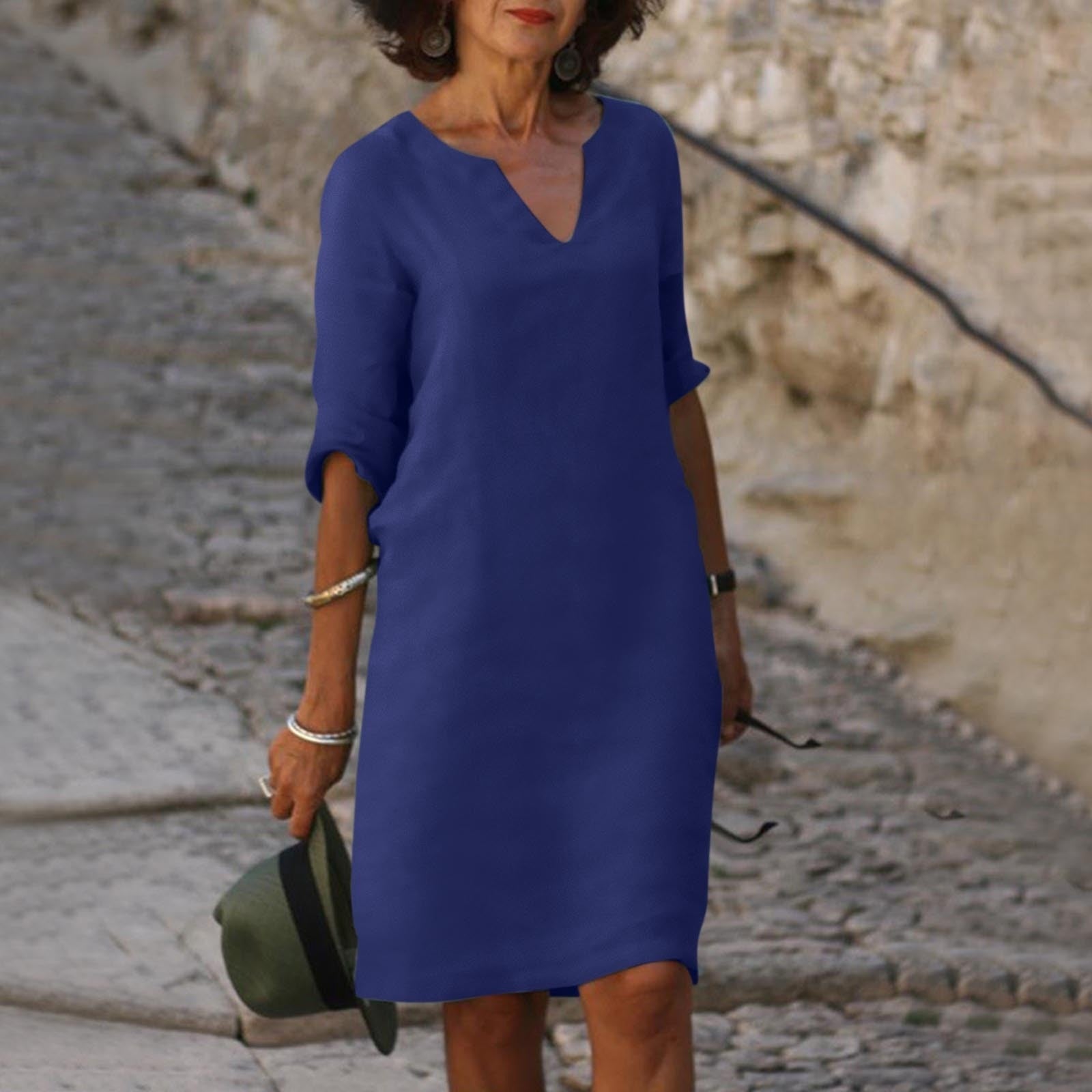 Donna con abito blu casual, maniche a tre quarti, su strada acciottolata. Moda estiva elegante, accessori argento, cappello verde in mano.