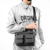 Borsa a tracolla nera unisex, design moderno con doppie tasche frontali, indossata su giacca grigia. Accessorio moda casual e funzionale.