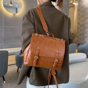 Zaino in pelle marrone elegante, indossato da una persona in giacca scura, in un ambiente moderno. Design con fibbie e dettagli dorati.