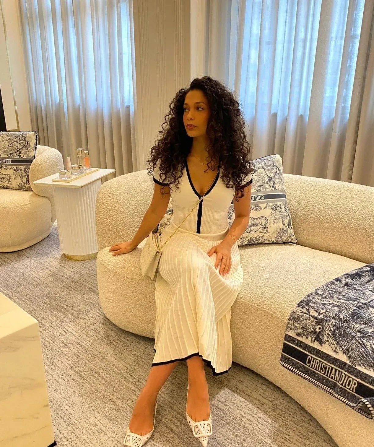 Donna con abito bianco seduta su divano elegante, interni lussuosi, borsa a tracolla, scarpe bianche, arredamento moderno, tenda chiara.