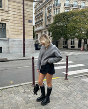 Donna con maglione grigio oversize, minigonna nera e stivali alti neri in strada cittadina. Moda autunnale urbana, look casual chic.