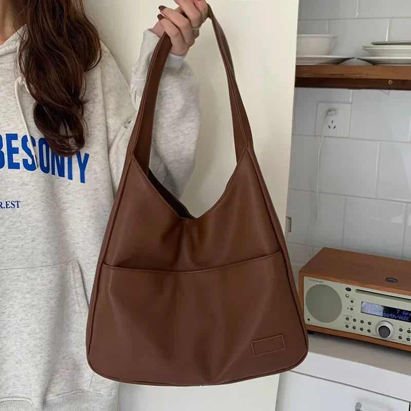 Donna con borsa a spalla in pelle marrone, design minimalista, indossa felpa grigia. Accessorio moda elegante, perfetto per uso quotidiano.