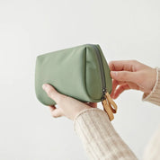 Mani che tengono un astuccio verde in pelle con cerniera dorata, sfondo bianco. Accessorio moda, borsa trucco elegante, design minimalista.
